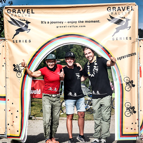Drei Teammitglieder der Gravel Rallye Black Forest posieren lächelnd unter dem bunten Startbogen.